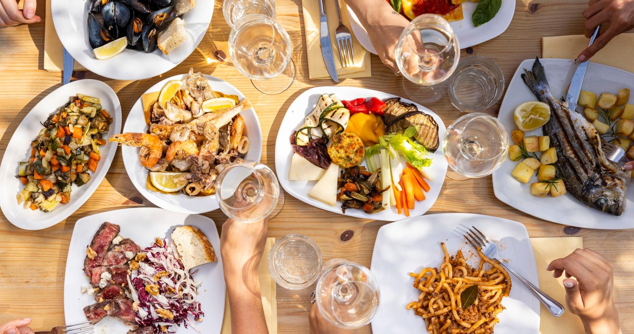 Vista dall'alto di una tavola imbandita con diversi piatti: insalata e verdure, pesce, carne, patatine fritte, pane e crostini, accompagnati da bicchieri di vino e acqua; mani di commensali intorno al tavolo.