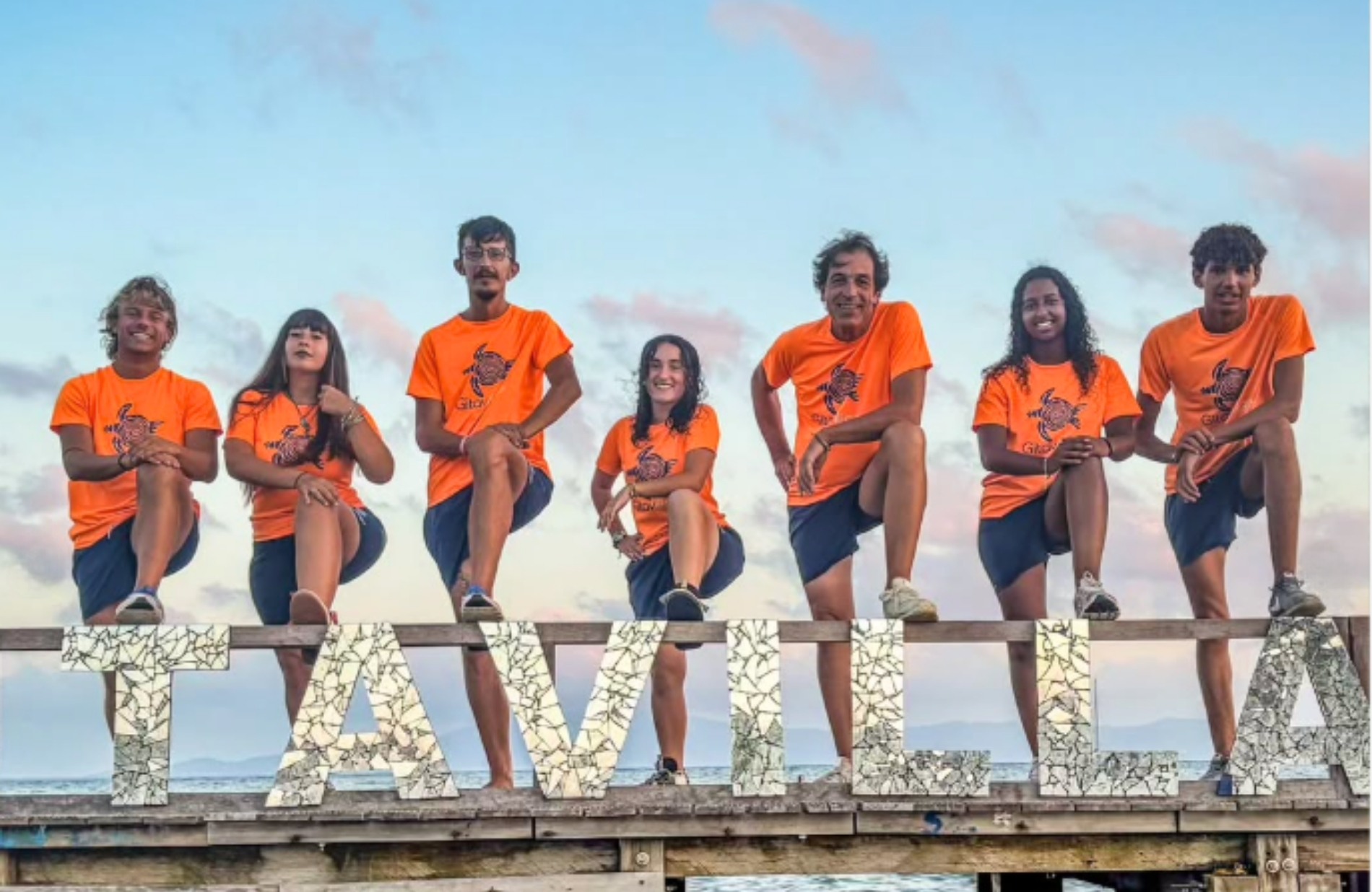 Un gruppo di otto persone in magliette arancioni in piedi su una ringhiera di legno, in equilibrio sopra grandi lettere mosaicate che formano la parola TAVILLA, con il cielo azzurro sullo sfondo.