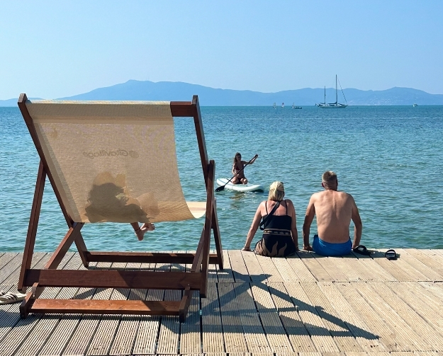 Ein Holzsteg am Strand mit einer großen Strandliege links. Zwei Personen sitzen am Rand des Stegs und schauen aufs Meer; im Wasser paddelt eine Person auf einem Stand-Up-Paddleboard. In der Ferne liegt ein Segelboot, und im Hintergrund ziehen sich Bergketten über den klaren blauen Himmel.