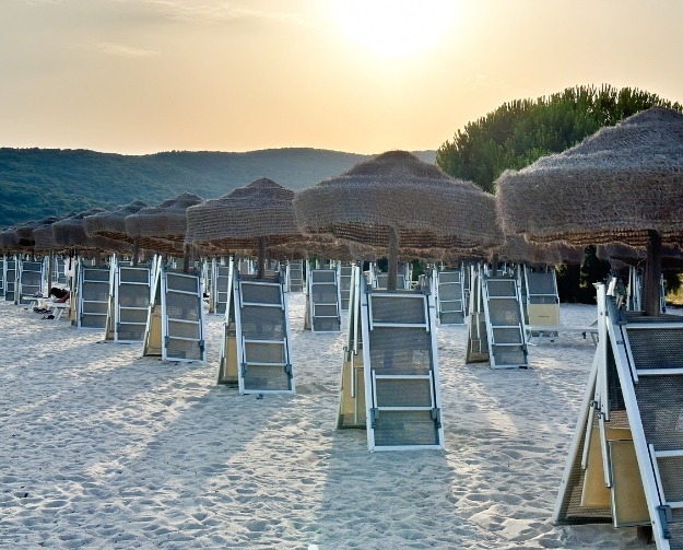 Ein Strand mit zahlreichen Strohsonnenschirmen und Liegen im Sand, bei Sonnenuntergang.