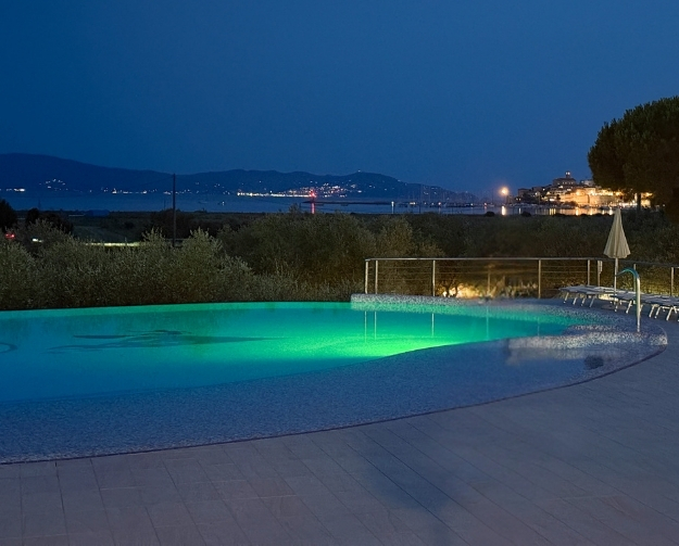 Beleuchteter Außenpool bei Nacht mit grünem Wasser; die flache, runde Kante besteht aus Fliesen, im Hintergrund sind Büsche und eine Küstenlandschaft mit Lichtern zu sehen, rechts stehen Liegen und ein Sonnenschirm am Beckenrand.