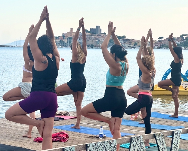 Eine Gruppe von Menschen macht Yoga-Übungen auf einem Holzsteg am Wasser. Sie stehen in der Baum-Pose auf einem Bein, die Hände über dem Kopf zusammengeführt. Im Hintergrund sieht man das Meer und eine Küstenstadt.