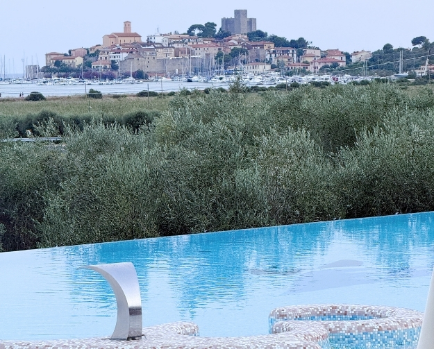Blick auf eine Küstenstadt auf einem Hügel mit Burganlage; im Vordergrund ein Infinity-Pool mit blauem Wasser, davor grüne Sträucher, dahinter ein Hafen mit Booten.